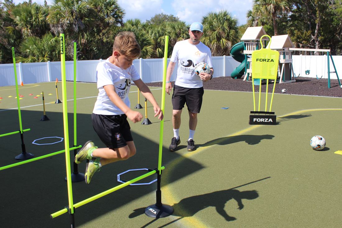 A student hops through agility poles while a coach looks on.