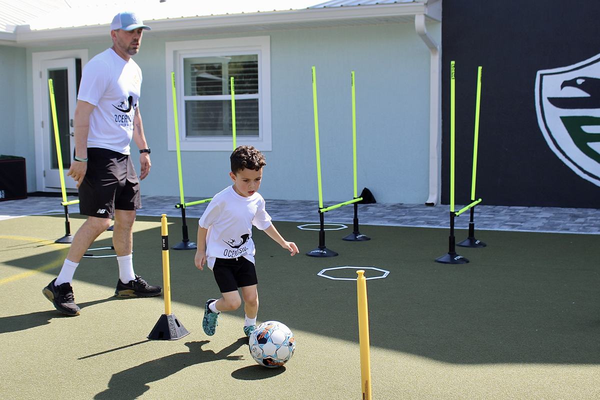 A coach jogs alongside a young student dribbling through training poles.