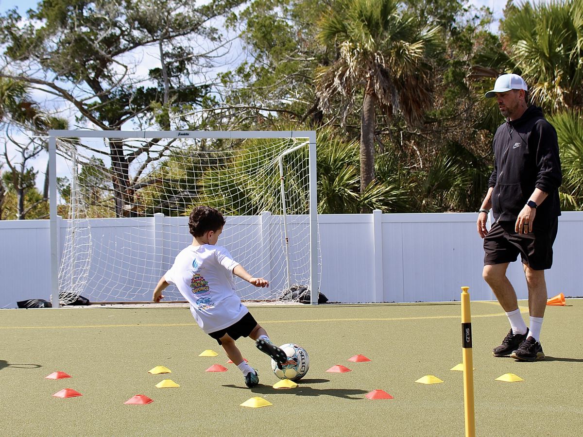 A coach watches a student dribble through a cone drill toward the goal.