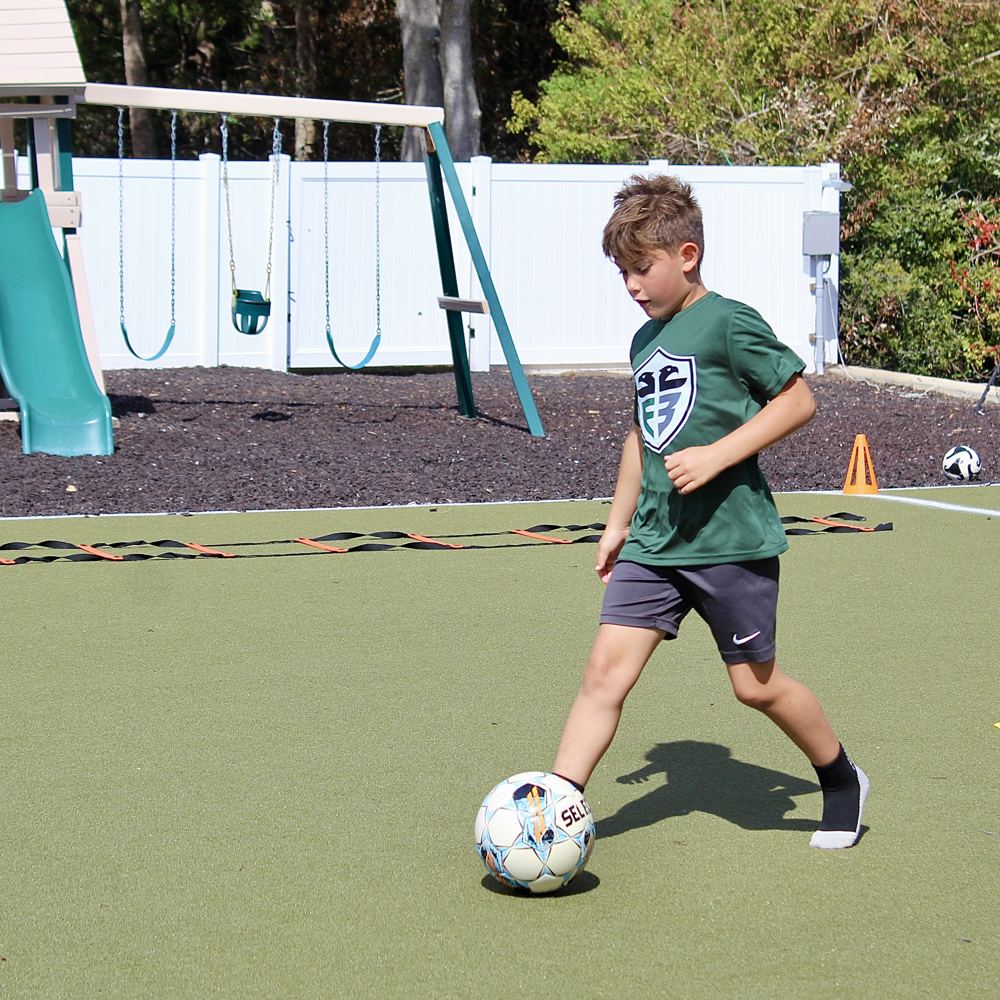 A student in a green jersey controls a soccer ball on the turf.