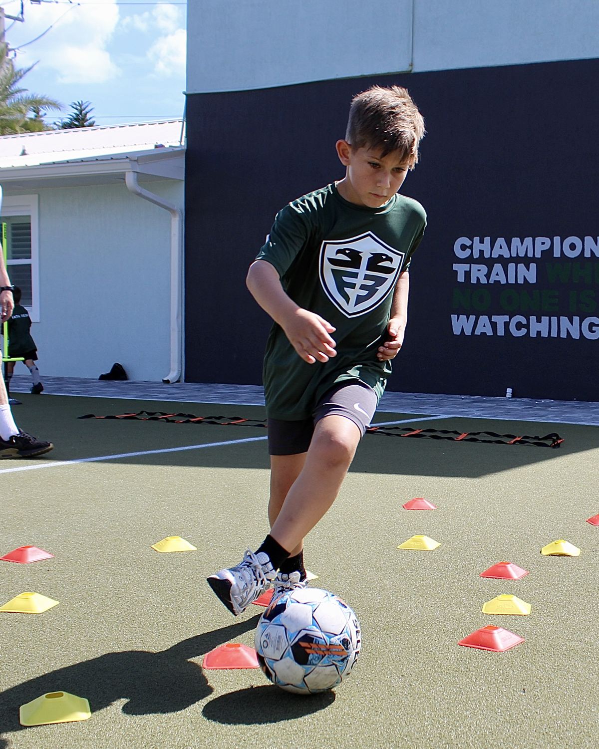 A young student in a green Faith & Freedom jersey dribbles a soccer ball through cones in front of the academy's training wall.