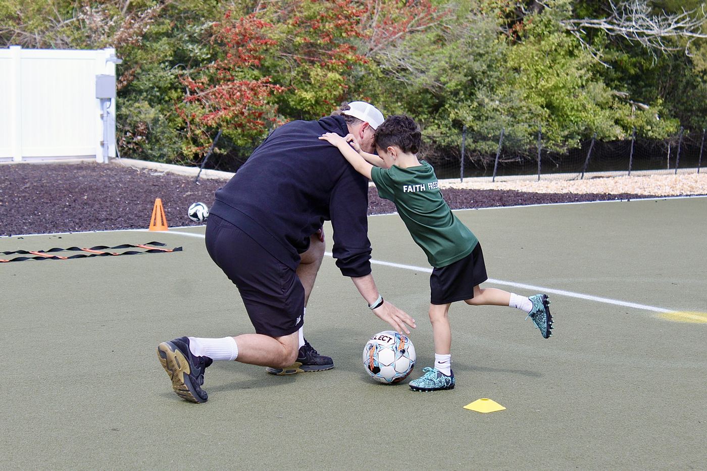 A coach kneels on the turf to show a student how to shield the ball.