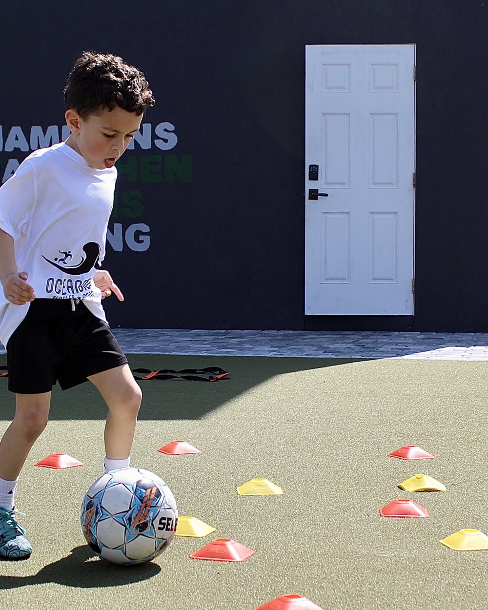 A young student dribbles through cones in front of the academy's training wall.
